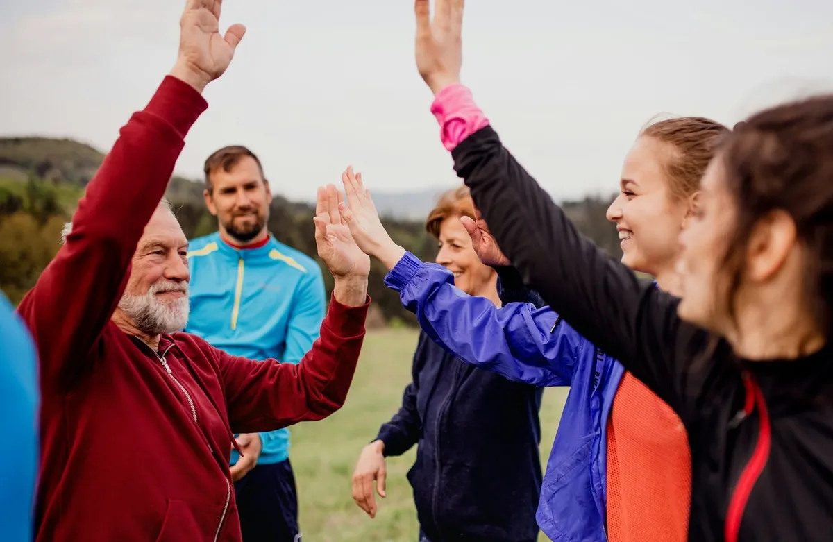 Oameni de vârste diferite, în haine de sport, bat palma cu mâinile ridicate, în aer liber