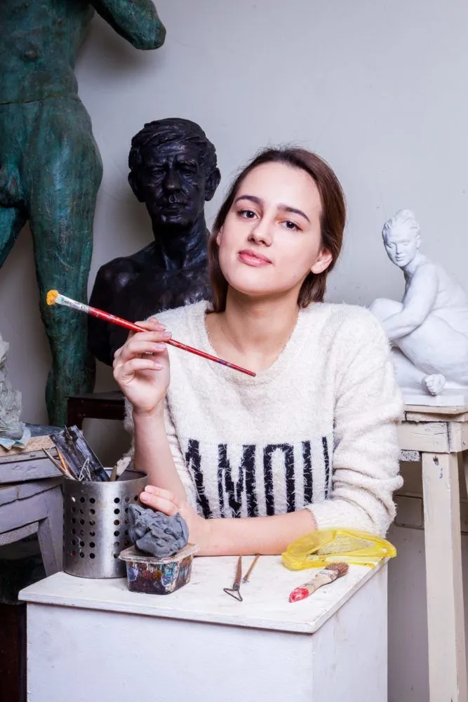 O tânără sprijinită de un postament alb, într-un atelier de sculptură, ține în mână o pensulă; în spatele ei, un bust de bronz, o statuie verde și o sculptură albă