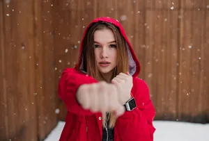 O tânără în geacă roșie cu glugă, cu un ceas inteligent la mână, întinde pumnul spre obiectiv, pe ninsoare