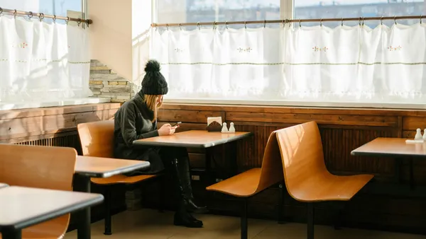 O tânără cu căciulă neagră stă singură la o masă într-o cafenea luminoasă și se uită în telefon