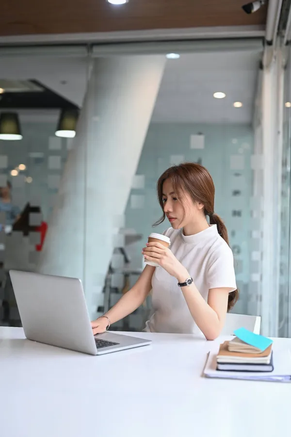 O tânără cu un pahar de cafea în mână lucrează la laptop, la o masă albă, într-un birou luminos