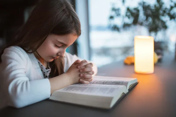 O fetiță se roagă cu mâinile împreunate peste o Biblie deschisă, lângă o lumânare aprinsă