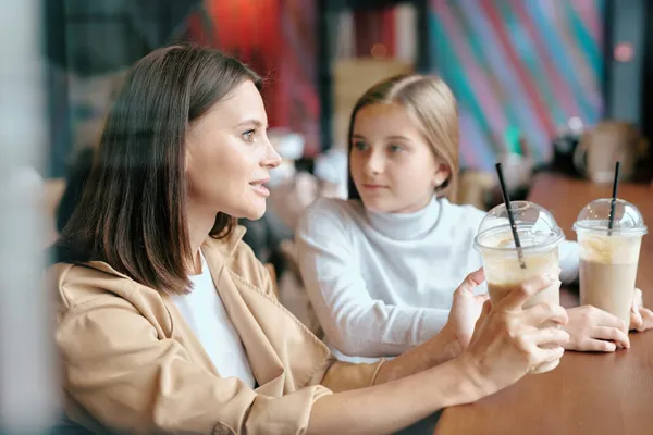 O mamă stă de vorbă cu fiica ei la o masă dintr-o cafenea, în fața a două pahare cu milkshake și pai