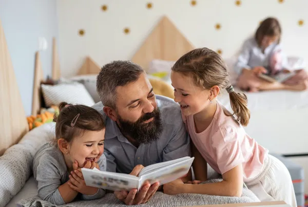 Un tată cu barbă, întins pe un pat, citește o carte cu două fetițe care râd; în fundal, o a treia fetiță citește singură