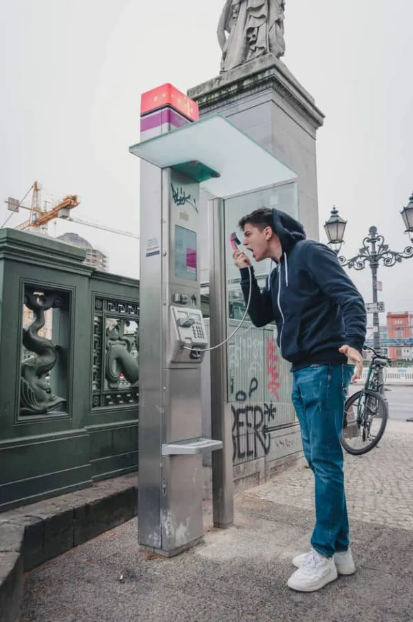 Un tânăr cu hanorac închis la culoare strigă în receptorul unui telefon public, pe un trotuar dintr-un oraș