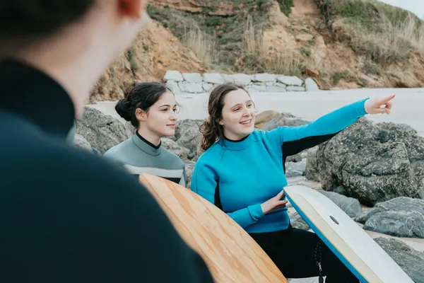 Două tinere în costume de neopren, cu plăci de surf, stau pe o plajă cu stânci; una arată cu mâna în depărtare, iar în prim-plan, din spate, cineva le ascultă