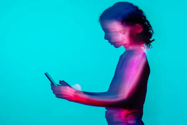 O fată cu părul creț, fotografiată din profil pe un fundal turcoaz, se uită la telefonul din mâini, cu imaginea estompată de mișcare și lumină roz