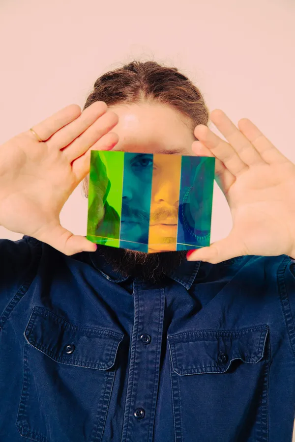 Un bărbat cu barbă și cămașă de blugi ține în fața feței o prismă colorată, prin care chipul lui apare împărțit în fâșii verzi, albastre și portocalii