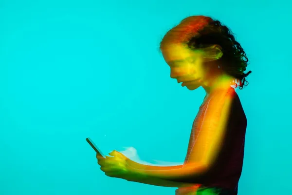 O adolescentă cu părul creț se uită la telefon, din profil, într-o fotografie cu lumini colorate și efect de mișcare, pe fundal turcoaz