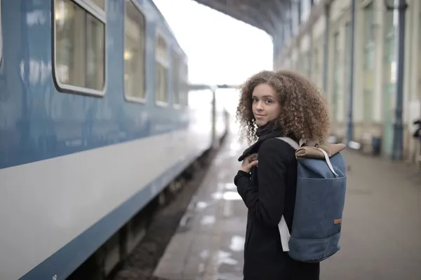 O tânără cu părul creț și un rucsac albastru stă pe peronul unei gări, lângă un tren albastru, și privește peste umăr