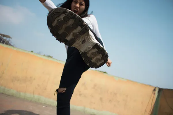 O tânără cu gura deschisă, ca și cum ar striga, ridică piciorul spre aparatul de fotografiat; în prim-plan se vede talpa pantofului ei sport