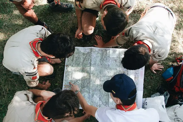 Cercetași în cămăși bej, cu eșarfe roșii, aplecați pe iarbă în jurul unei hărți