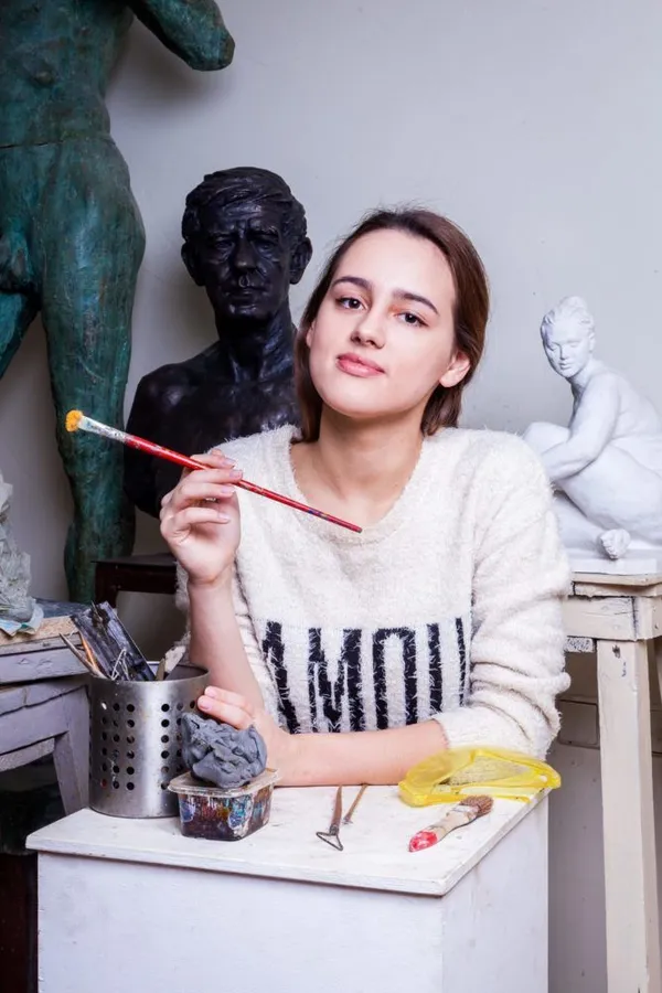 O tânără sprijinită de un postament alb, într-un atelier de sculptură, ține în mână o pensulă; în spatele ei, un bust de bronz, o statuie verde și o sculptură albă