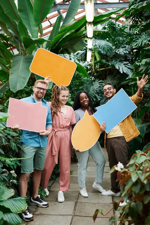 Patru tineri zâmbitori, într-o seră plină de plante tropicale, țin în mâini baloane de dialog colorate din carton, fără text